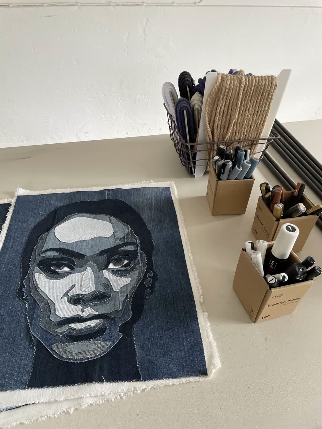 A human face pieced from recycled indigo denim beside a workshop shelf of rope, cord and markers.