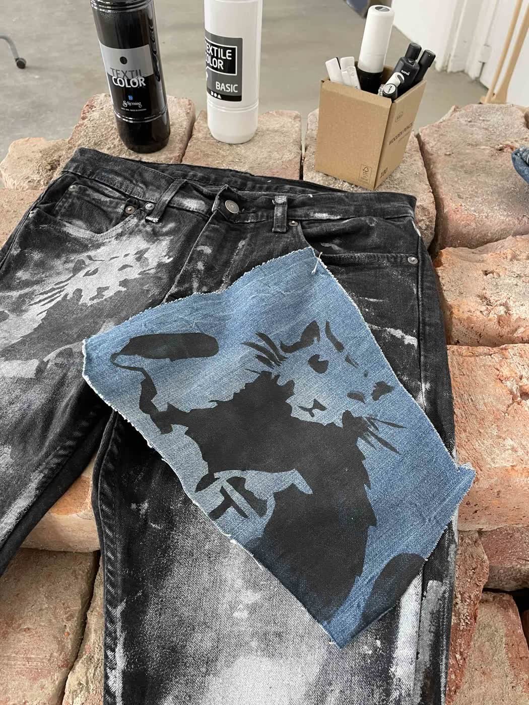 Close-up of a raw-edge blue denim patch printed with a punk cat graphic on bleach-splattered black jeans.