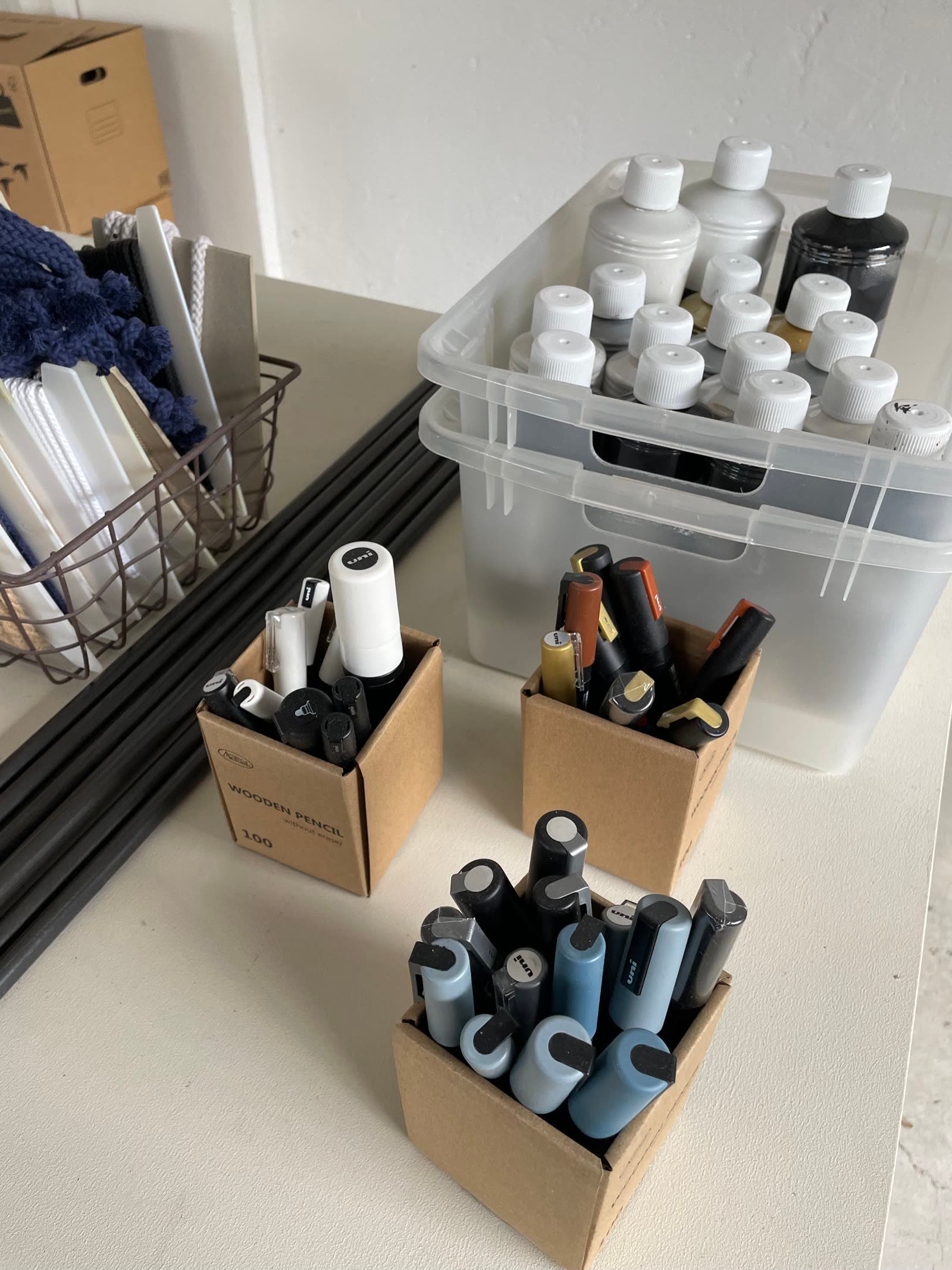 Boxes of textile-paint bottles and marker pens in a tub on the workshop table.